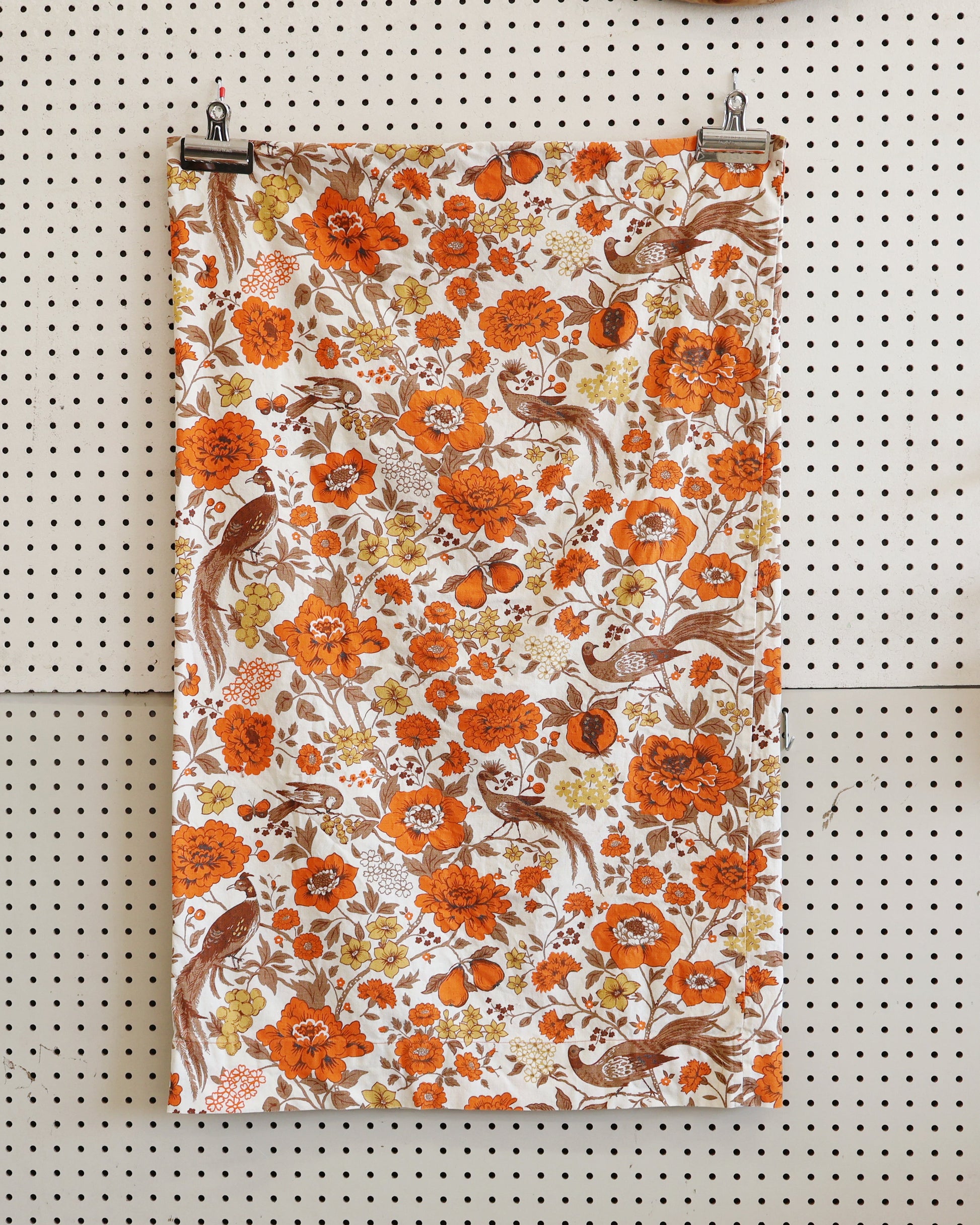 A vertical fabric panel or poster print featuring a vintage-style floral and bird pattern in orange, brown, and cream, hanging from two black clips on a white pegboard wall.